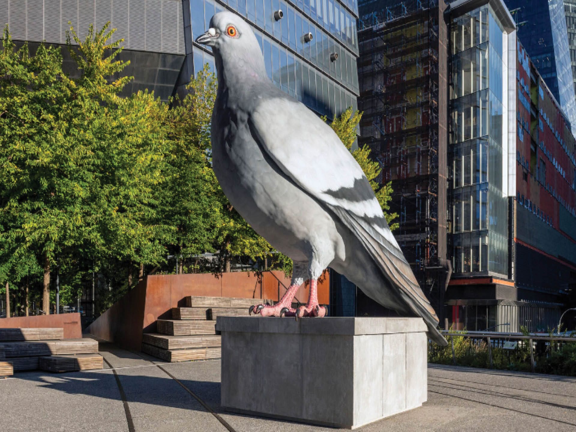 A giant pigeon sculpture stands proudly on a concrete pedestal along the High Line, surrounded by modern buildings and lush greenery. The artwork is intricately detailed, depicting the bird with lifelike features, poised as if observing its vibrant urban surroundings.