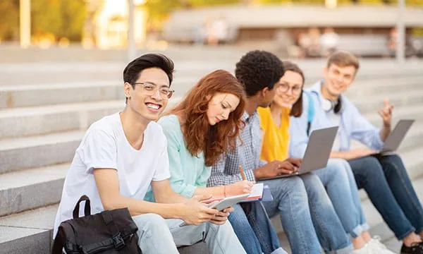 A group of five young adults sitting on outdoor steps, studying and chatting. One person in the foreground smiles at the camera, holding a notebook. Others are focused on laptops and taking notes. The background is sunny with blurred trees.