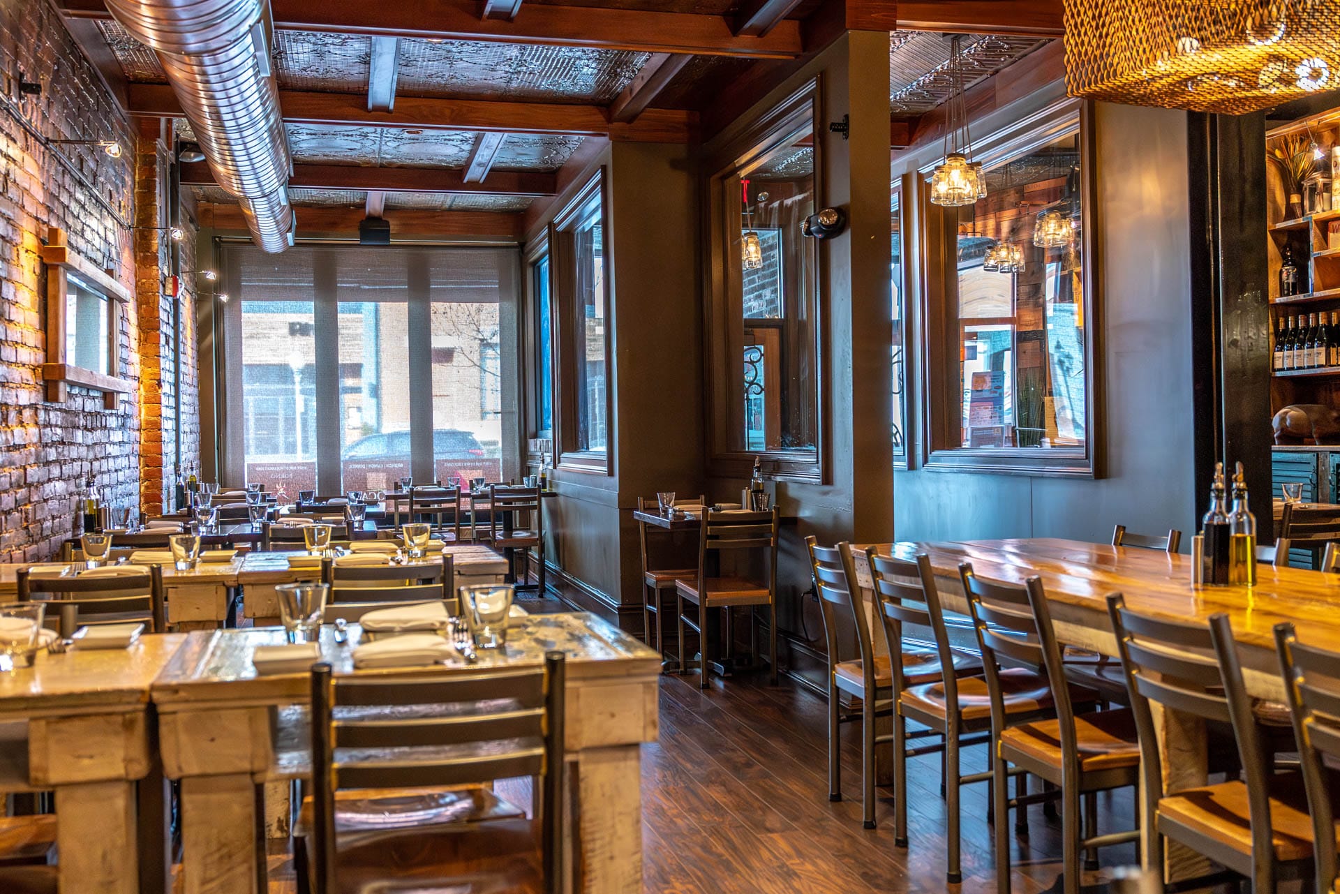 Warm, cozy interior at Boca Bistro with wooden tables and chairs, exposed brick walls, and rustic decor. Natural light filters through large windows, creating a welcoming atmosphere. Tables are elegantly set with glassware and cutlery.