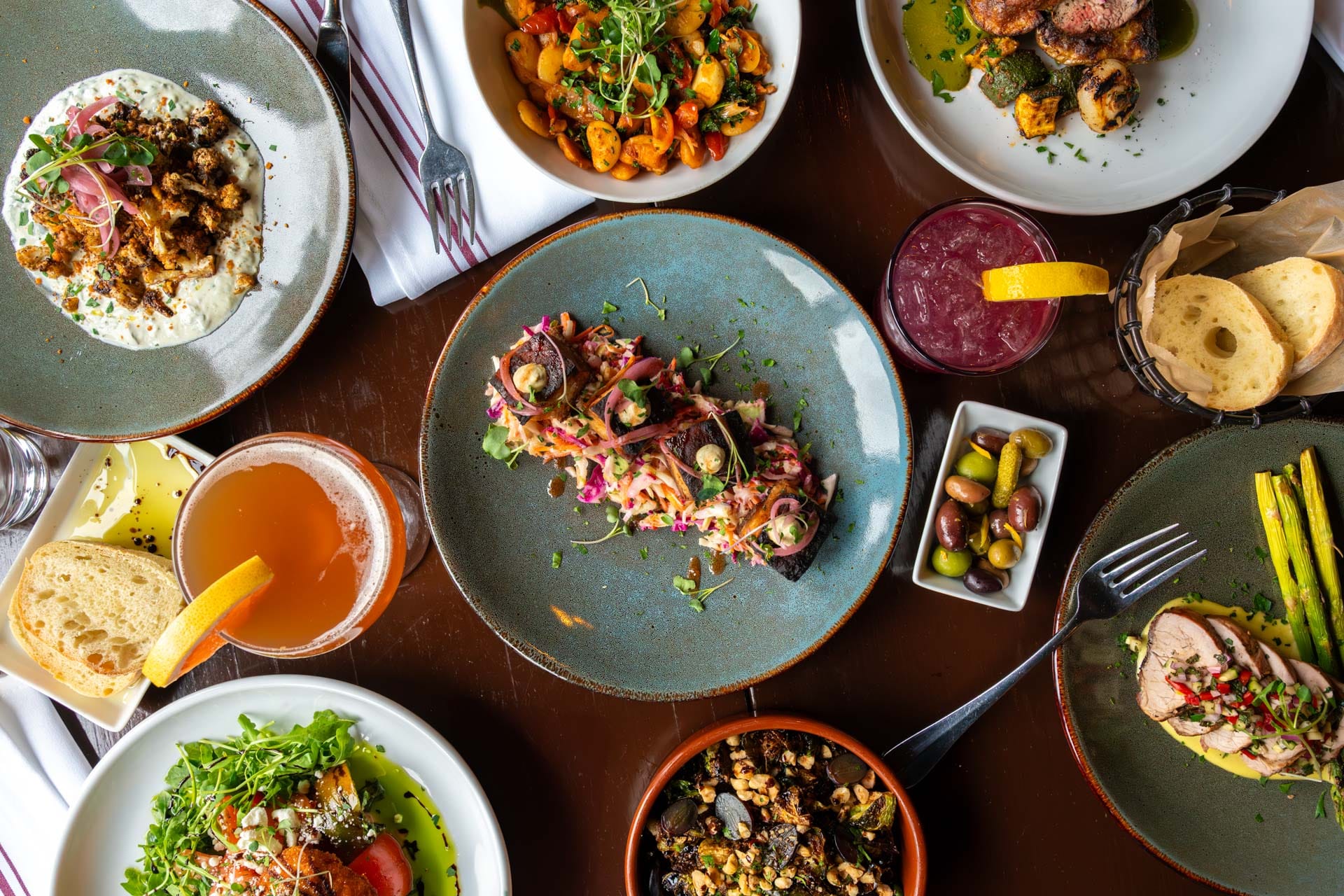 An overhead view of a dining table at Boca Bistro showcases a colorful salad, bowls of mixed olives, a basket of sliced bread, roasted vegetables, grilled meat, and cocktails—all artfully arranged for an exquisite dining experience.