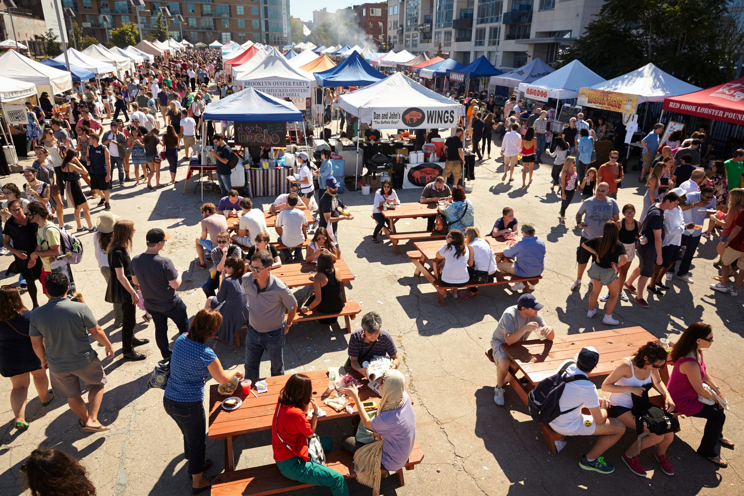 A bustling outdoor food festival with numerous white and colored tents. People are walking, socializing, and seated at picnic tables eating. The scene is lively with diverse attendees and vibrant stalls in a sunlit urban area.