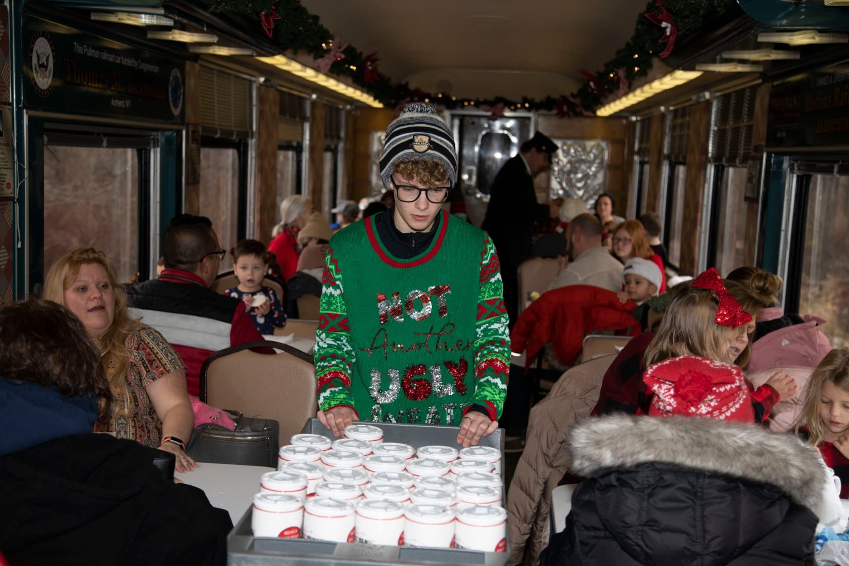 A person wearing a festive sweater and hat serves cups of hot chocolate on the Medina Railroad Polar Express, adorned with holiday garlands. Passengers, including children in Christmas attire, are seated at tables, reveling in the magical festive atmosphere.