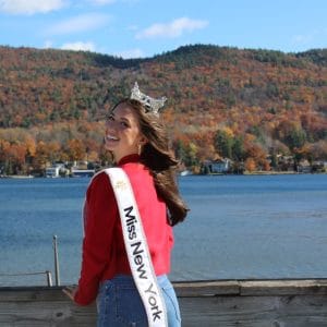 A woman in a red sweater and Miss New York sash smiles by Lake George, crowned with elegance. The background features autumn-colored trees and a hillside under the blue sky, capturing the serene beauty of this iconic location.